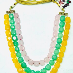 May include: A three-strand necklace with yellow, green, and pink beads. The beads are square-shaped and have a polished finish. The necklace has a gold clasp and a tassel.