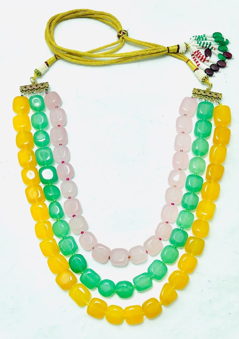 May include: A three-strand necklace with yellow, green, and pink beads. The beads are square-shaped and have a polished finish. The necklace has a gold clasp and a tassel.