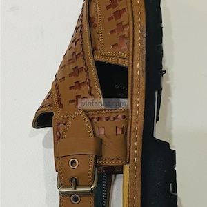 May include: Brown leather sandal with a woven design on the upper and a black sole. The sandal has a buckle closure and is handmade. The brand name "VintaRust" is visible.