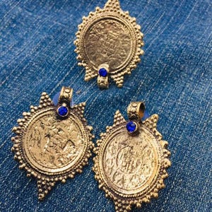 May include: Three gold-toned pendants with intricate designs and blue gemstone accents. Each pendant features a circular shape with a raised, textured surface.