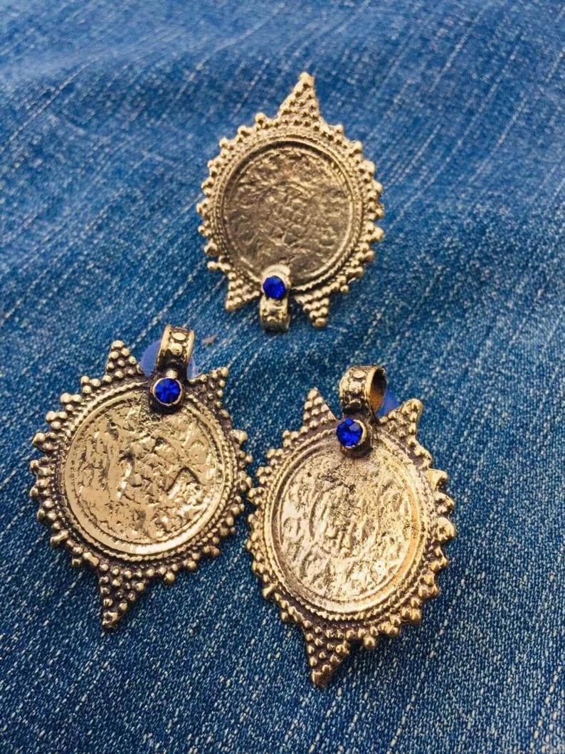 May include: Three gold-toned pendants with intricate designs and blue gemstone accents. Each pendant features a circular shape with a raised, textured surface.