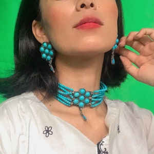 May include: A woman wearing a white shirt with black embroidery and a turquoise choker necklace with a pendant. She is also wearing turquoise earrings.