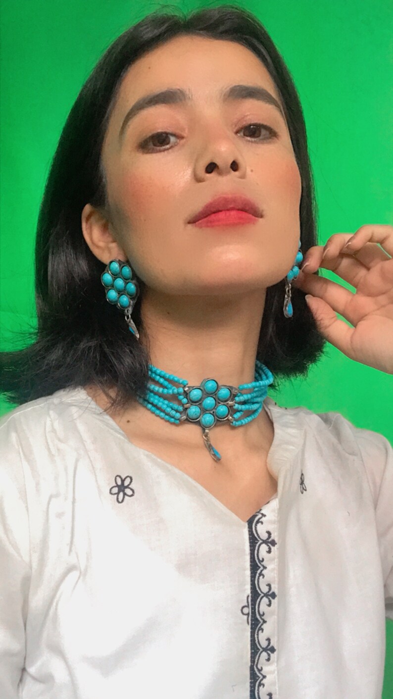May include: A woman wearing a white shirt with black embroidery and a turquoise choker necklace with a pendant. She is also wearing turquoise earrings.