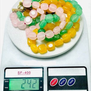 May include: A close-up of a digital scale displaying 242 grams. Three strands of beads are on the scale, one strand is yellow, one is green, and one is pink.
