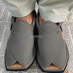 K&ouml;nnte beinhalten: Ein Paar graue Peshawari-Sandalen aus Leder mit goldfarbenen Zierleisten und einem hellrosa Akzent. Die Sandalen haben ein einzigartiges Design mit spitzer Zehenpartie und einem dekorativen Emblem. Handgefertigte Klassiker.