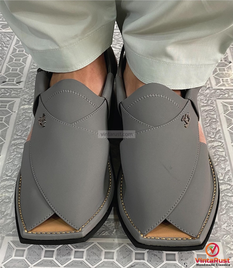 K&ouml;nnte beinhalten: Ein Paar graue Peshawari-Sandalen aus Leder mit goldfarbenen Zierleisten und einem hellrosa Akzent. Die Sandalen haben ein einzigartiges Design mit spitzer Zehenpartie und einem dekorativen Emblem. Handgefertigte Klassiker.