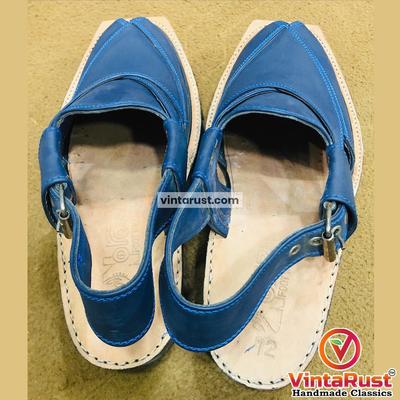 May include: A pair of blue leather sandals with a unique design. The sandals feature a square toe, stitched detailing, and adjustable straps with metal buckles. The soles are a light tan color, and the sandals appear to be handmade.