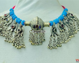 May include: A silver-toned necklace with a central ornate pendant and dangling charms. The necklace features a blue cord and decorative elements with red and blue accents. The necklace is displayed on a white surface.