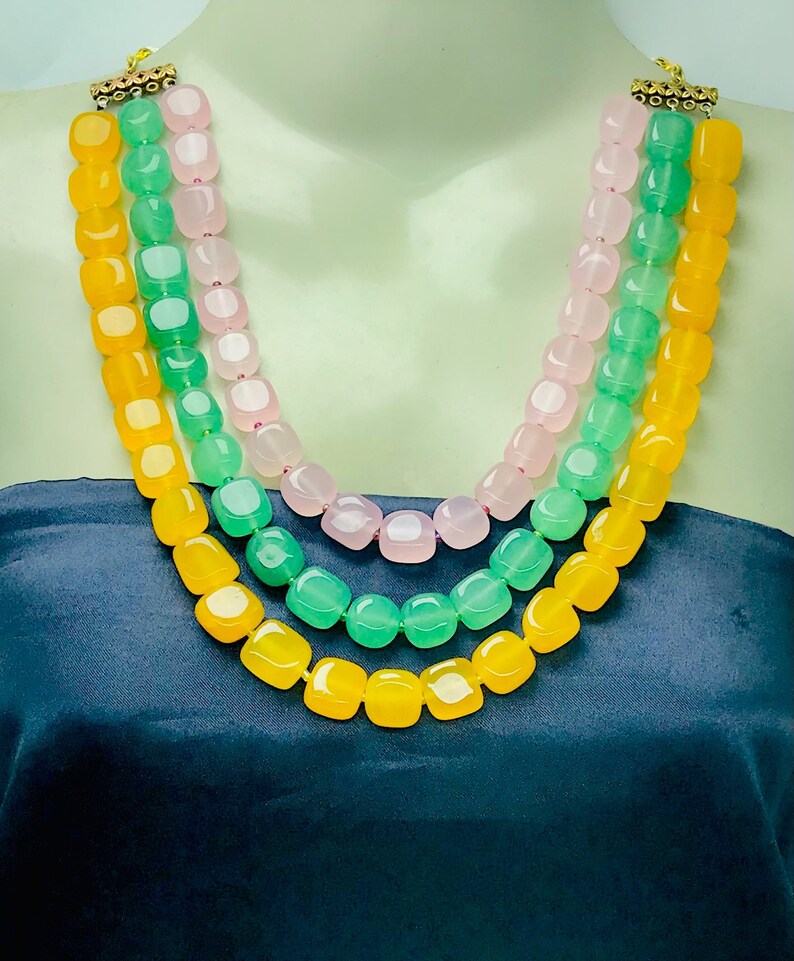 May include: A three-strand necklace with yellow, green, and pink square beads. The necklace is made of semi-precious stones and has a gold clasp.