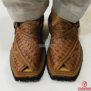 May include: A pair of brown leather Peshawari sandals with a woven design. The sandals have a square toe, a strap around the ankle, and a black sole. The image shows the sandals from a top-down perspective.