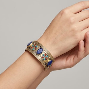 May include: A wide gold-tone metal cuff bracelet featuring an intricate engraved pattern, inlaid with oval blue lapis lazuli stones and small flower-shaped clusters of turquoise and red coral-colored cabochons arranged along the center band.