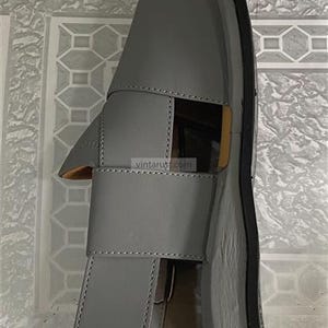 K&ouml;nnte beinhalten: Graue Ledersandale mit einem Design mit einem einzigen Riemen. Die Sandale hat eine dicke Sohle und eine minimalistische &Auml;sthetik. Die Sandale ist etwa 25 cm lang.