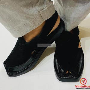 May include: Black leather Peshawari sandals with a glossy toe cap and a suede upper. The sandals have a traditional design with a strap around the heel. The footwear is handmade and suitable for casual wear.