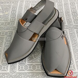 K&ouml;nnte beinhalten: Ein Paar graue Ledersandalen mit Riemen und Schnallenverschluss. Die Sandalen haben ein gen&auml;htes Design an den Kanten und eine hellbraune Innenseite. Die Sohlen sind schwarz mit einer hellgrauen Zwischensohle. Die Sandalen werden auf einer gemusterten Oberfl&auml;che pr&auml;sentiert.