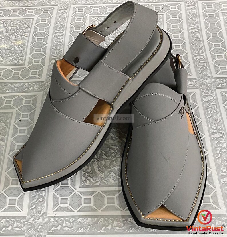 K&ouml;nnte beinhalten: Ein Paar graue Ledersandalen mit Riemen und Schnallenverschluss. Die Sandalen haben ein gen&auml;htes Design an den Kanten und eine hellbraune Innenseite. Die Sohlen sind schwarz mit einer hellgrauen Zwischensohle. Die Sandalen werden auf einer gemusterten Oberfl&auml;che pr&auml;sentiert.
