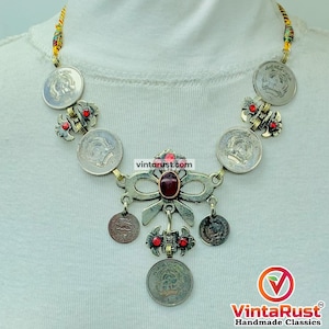 May include: A silver-toned necklace featuring coin pendants and decorative elements. The necklace has a central bow-shaped pendant with a red stone and smaller red accents. The cord is multi-colored.