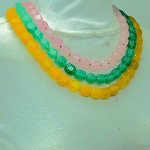 May include: Three strands of beads in a necklace. The top strand is pink, the middle strand is green, and the bottom strand is yellow. The beads are all square shaped.