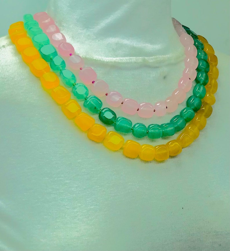 May include: Three strands of beads in a necklace. The top strand is pink, the middle strand is green, and the bottom strand is yellow. The beads are all square shaped.