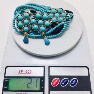 May include: A turquoise and brass necklace with multiple strands of beads and a pendant. The necklace is on a digital scale that reads 21 grams.