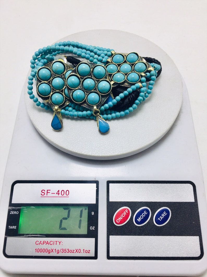 May include: A turquoise and brass necklace with multiple strands of beads and a pendant. The necklace is on a digital scale that reads 21 grams.