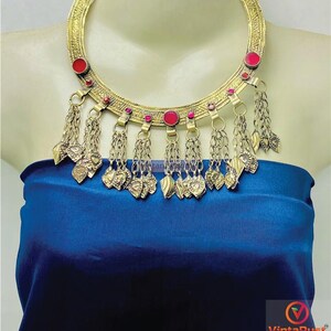Può includere: Una collana color oro con accenti rossi e ciondoli pendenti. La collana presenta un ampio colletto con pietre rosse circolari e accenti rossi più piccoli. Catene con ciondoli a forma di cuore e foglia pendono dal colletto. La collana è esposta su un manichino.