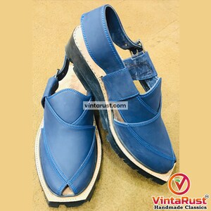 May include: A pair of blue leather Peshawari sandals with a black sole and beige trim. The sandals have a strap and buckle closure. The image includes the text "vintarust.com" and the VintaRust Handmade Classics logo.