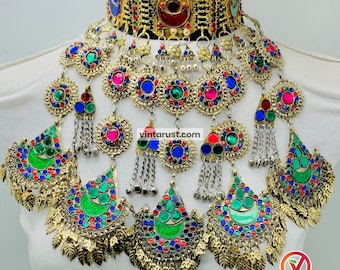 May include: A multi-layered gold necklace with colorful glass stones and silver chains. The necklace features a large, round pendant with a crescent moon design and a smaller pendant with a floral design. The necklace is adorned with tassels and bells.