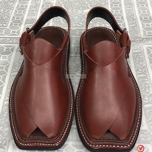 Puede incluir: Un par de sandalias Peshawari de cuero marr&oacute;n. Presentan una puntera cuadrada, una correa en el tobillo y un cierre de hebilla. El cuero es liso y los bordes est&aacute;n cosidos con hilo blanco. Dise&ntilde;o tradicional.