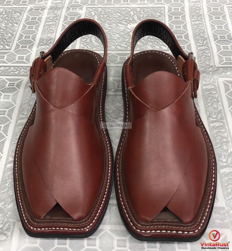 Puede incluir: Un par de sandalias Peshawari de cuero marr&oacute;n. Presentan una puntera cuadrada, una correa en el tobillo y un cierre de hebilla. El cuero es liso y los bordes est&aacute;n cosidos con hilo blanco. Dise&ntilde;o tradicional.
