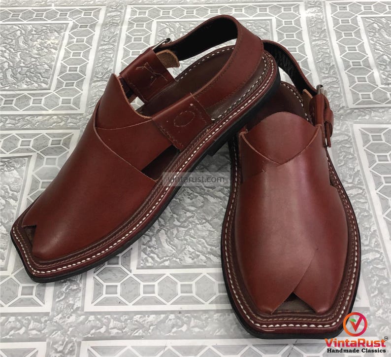 Puede incluir: Un par de sandalias de cuero marr&oacute;n con un dise&ntilde;o distintivo. Las sandalias tienen una correa en la parte superior y un cierre de hebilla. Las costuras blancas acent&uacute;an los bordes. Las sandalias se muestran sobre una superficie estampada.