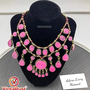 May include: A pink and gold statement necklace with a large round pendant and multiple teardrop-shaped stones. The necklace is made of metal and features a layered design with smaller beads.