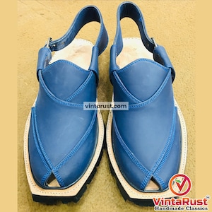 May include: A pair of blue leather Peshawari sandals. The sandals have a strap that goes around the ankle and a buckle closure. The soles are black and the stitching is blue. The website vintarust.com is visible on the sandals.