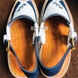 Pu&ograve; includere: Un paio di scarpe in pelle bianca con finiture nere e una fibbia dorata. Le scarpe hanno il numero "804" scritto su di esse.