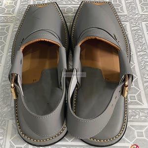 K&ouml;nnte beinhalten: Ein Paar graue Peshawari-Sandalen aus Leder mit quadratischer Zehenpartie. Die Sandalen haben einen Riemen mit einer goldfarbenen Schnalle und sind mit goldfarbenem Faden an den R&auml;ndern gen&auml;ht. Die Innenseite ist hellbraun.