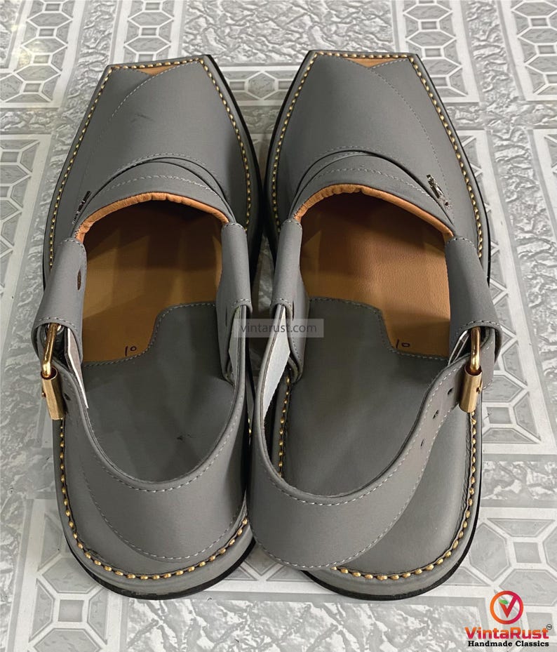 K&ouml;nnte beinhalten: Ein Paar graue Peshawari-Sandalen aus Leder mit quadratischer Zehenpartie. Die Sandalen haben einen Riemen mit einer goldfarbenen Schnalle und sind mit goldfarbenem Faden an den R&auml;ndern gen&auml;ht. Die Innenseite ist hellbraun.