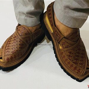 May include: Brown leather Peshawari sandals with a woven design. The sandals have a square toe and a black sole. The image shows the sandals being worn. The sandals are a traditional style of footwear.
