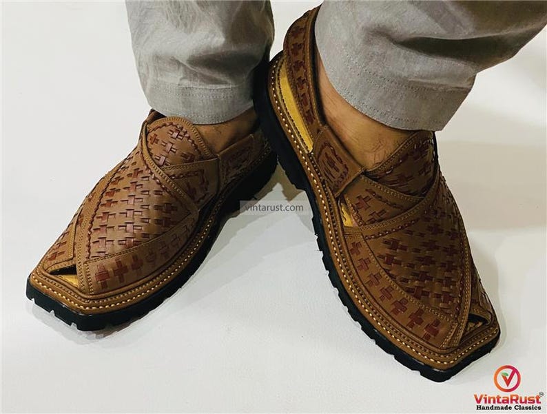 May include: Brown leather Peshawari sandals with a woven design. The sandals have a square toe and a black sole. The image shows the sandals being worn. The sandals are a traditional style of footwear.