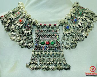 May include: An ornate silver necklace with colorful gemstone accents. The necklace features a central rectangular pendant and elaborate side pieces with dangling charms. The design incorporates red, green, and blue stones.
