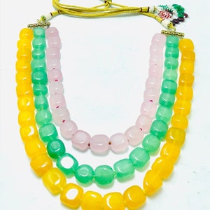 May include: A three-strand necklace with yellow, green, and pink square beads. The necklace has a gold clasp and a yellow cord.