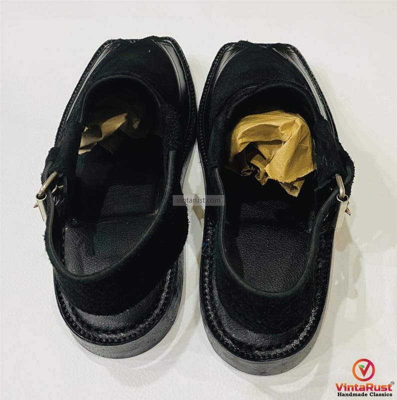 May include: A pair of black leather Peshawari sandals. These open-toe shoes feature a suede upper, a strap with a buckle closure, and a thick sole. The sandals are designed with a traditional silhouette and are suitable for casual wear.