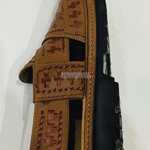 May include: A brown leather sandal with a woven design on the straps and upper. The sandal has a black sole and is likely handmade. The design includes cross-shaped stitching and a braided pattern. The sandal is a classic style.