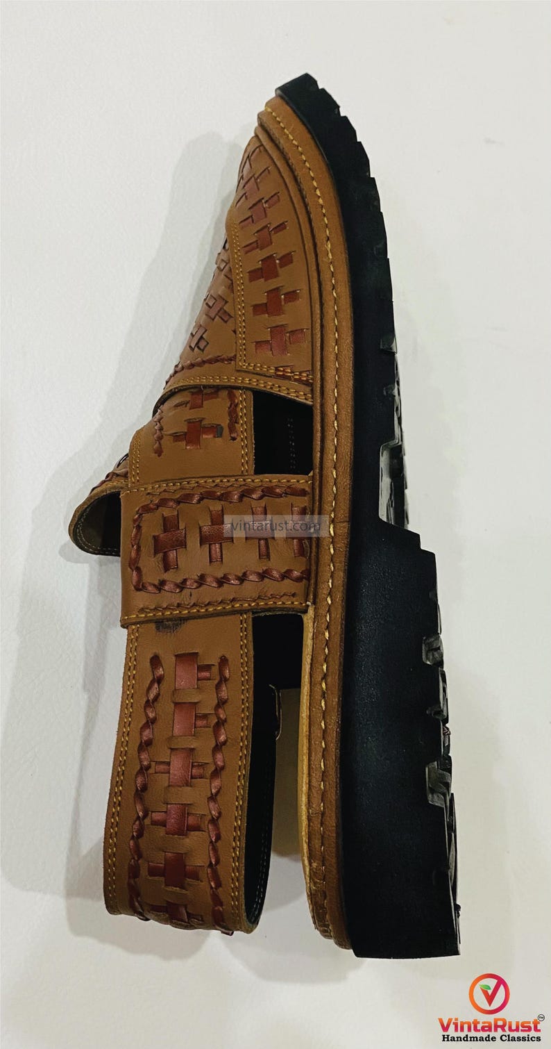 May include: A brown leather sandal with a woven design on the straps and upper. The sandal has a black sole and is likely handmade. The design includes cross-shaped stitching and a braided pattern. The sandal is a classic style.