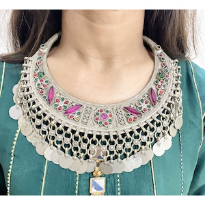 Può includere: Una collana d'argento con un ciondolo a forma di mezzaluna adornato con pietre preziose rosa e verdi. Il ciondolo è circondato da una catena di monete d'argento.