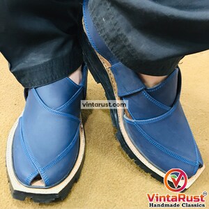 May include: Blue leather Peshawari sandals with light blue stitching. The sandals have a unique open-toe design and a thick, black sole. The brand name "VintaRust Handmade Classics" is visible in the bottom right corner.