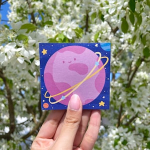 Può includere: Un pianeta rosa e viola con un viso carino e un anello giallo intorno. Il pianeta è circondato da stelle e uno sfondo blu.