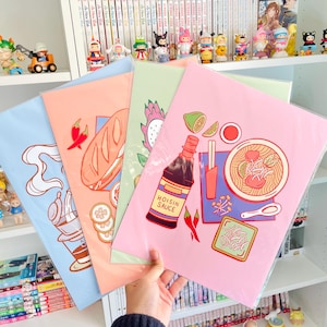 May include: A hand holds four colorful art prints. The prints feature illustrations of food items like a coffee cup, baguette, dragon fruit, and a bowl of noodles with the text "HOISIN SAUCE". The prints are on a white shelf.