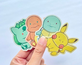 May include: Four cartoon character stickers featuring a green, yellow, blue, and orange character. The stickers are held in a hand.