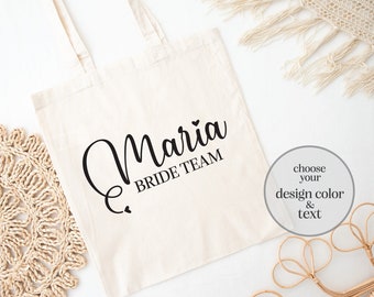 May include: A white canvas tote bag with black text that reads "Maria Bride Team". The bag is lying flat on a white surface with a woven straw mat and a piece of beige fabric in the background. A small circle with the text "choose your design color & text" is in the lower right corner of the image.