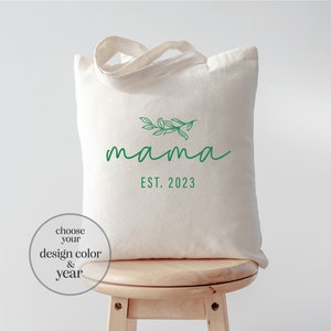 May include: A white canvas tote bag with a green leafy branch design and the text "mama est. 2023".
