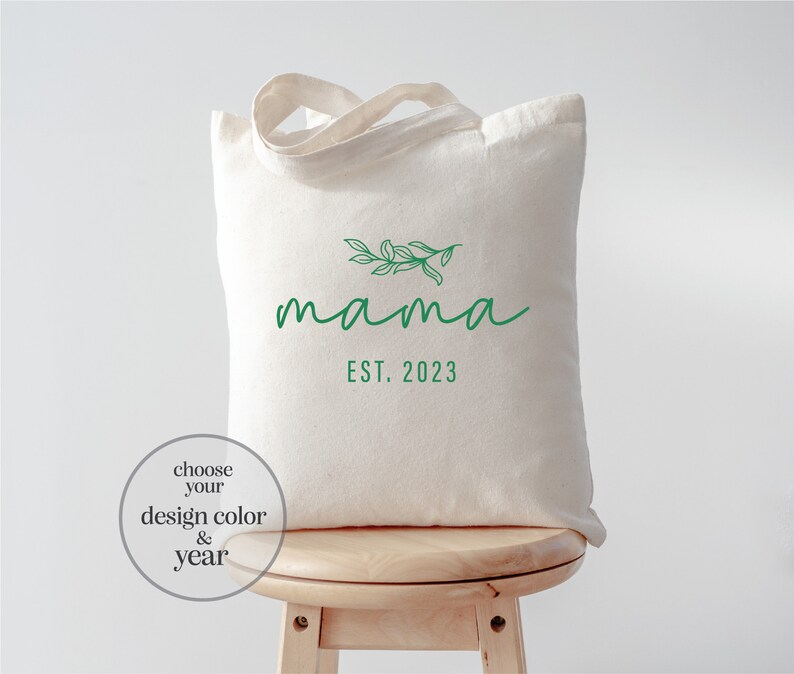 May include: A white canvas tote bag with a green leafy branch design and the text "mama est. 2023".